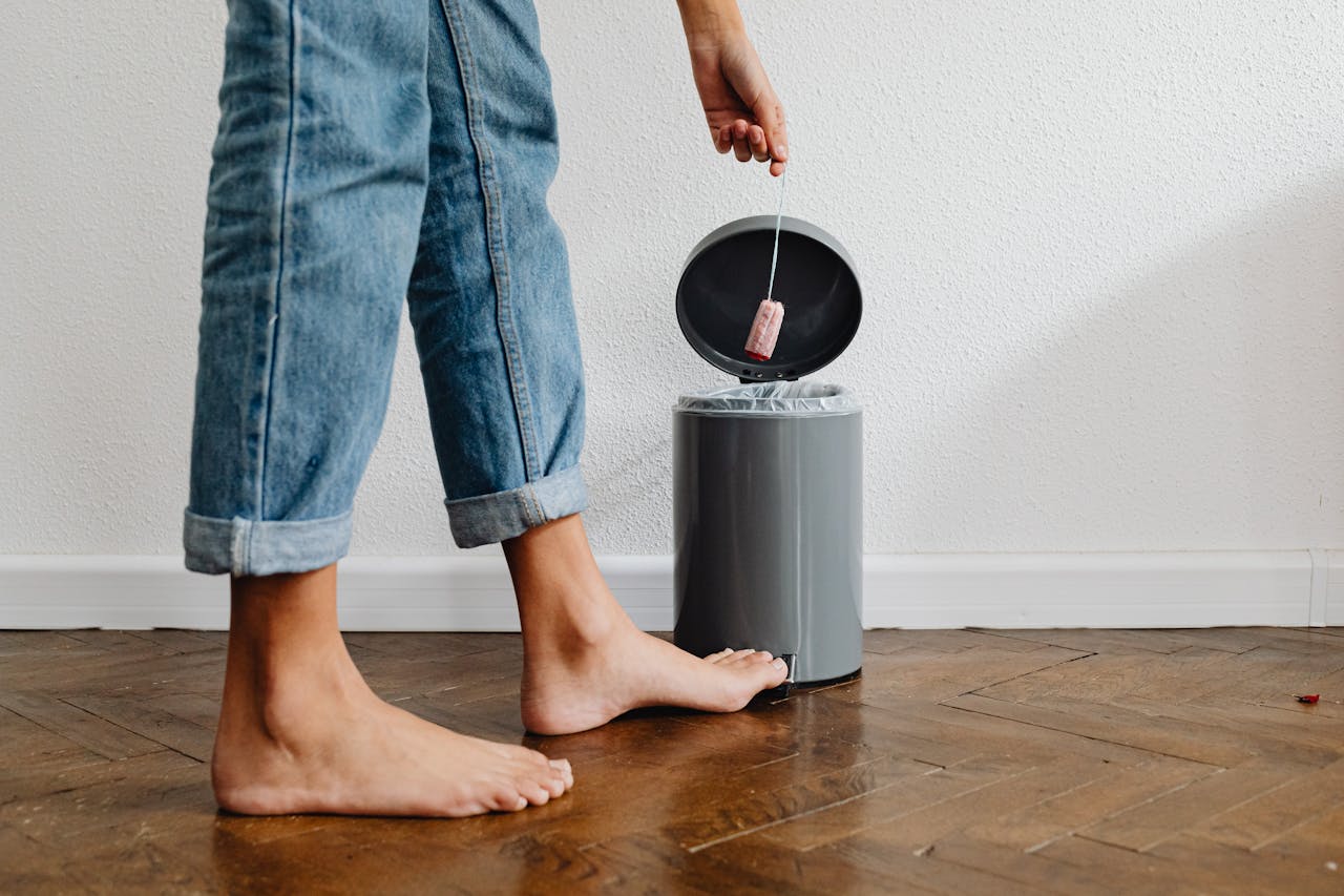 hero-img-01 Barefoot woman in blue jeans discarding a tampon in a trash can at home, symbolizing menstrual care.